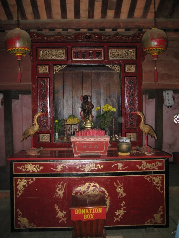 A shrine frame in red lacquer and gilt, four characters on the horizontal board above, a couplet running down the columns on each side, a small figure standing in the recess, two bronze cranes flanking the altar table, and two red lanterns hanging in front.