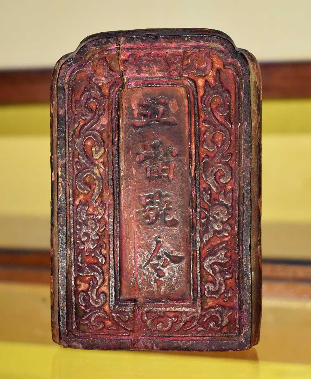 The front face of the tally: a block of wood in faded red lacquer, arched at the top, carved with a raised vine border, a rectangular panel at the centre cut with four characters in a vertical column, and a crack running down the piece.