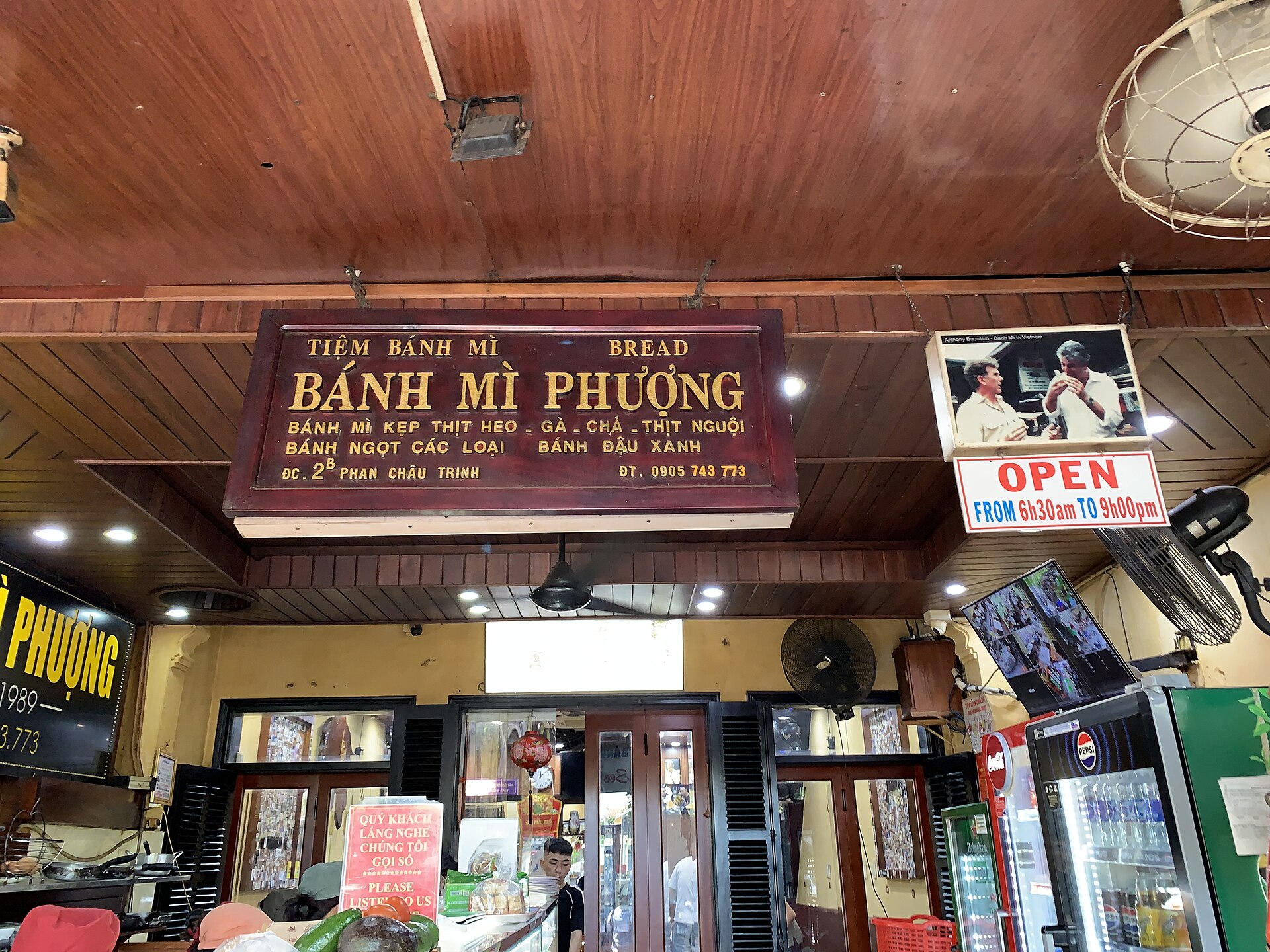 A wooden shop sign reading 'Tiệm bánh mì, Bánh mì Phượng' hangs above the door. To its right is a lit frame holding a photograph of two men eating bánh mì, with an English line above the photograph that names Anthony Bourdain.