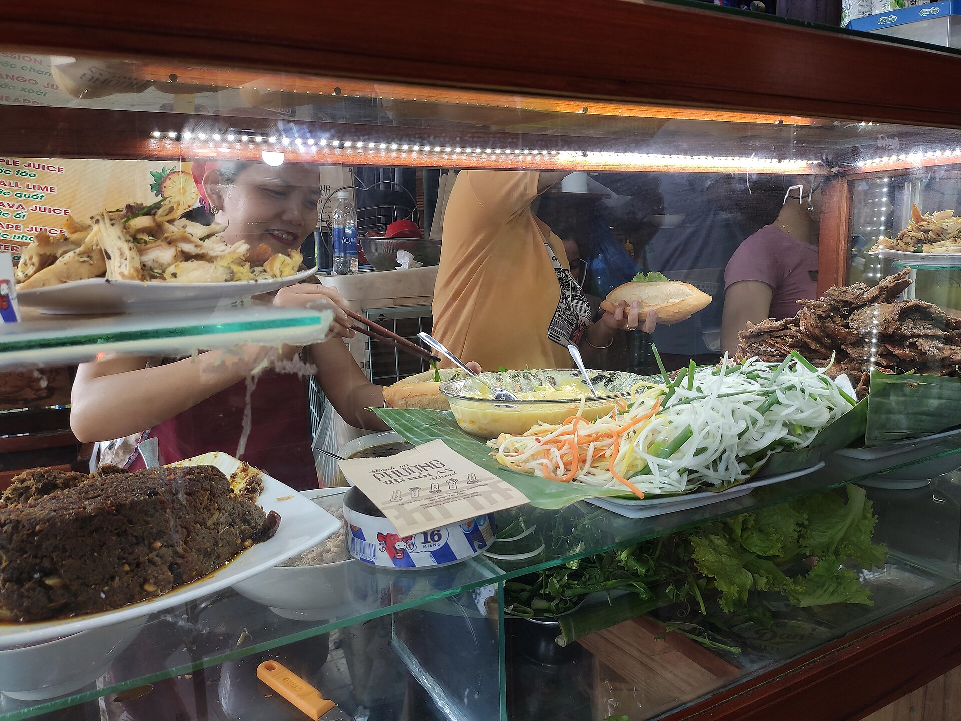 A glass-fronted bread stall seen from the front: a woman behind the counter lifts food with chopsticks, a bowl of egg and a mound of shredded onion and carrot on banana leaf sit in the middle, and one loaf lies beside them.