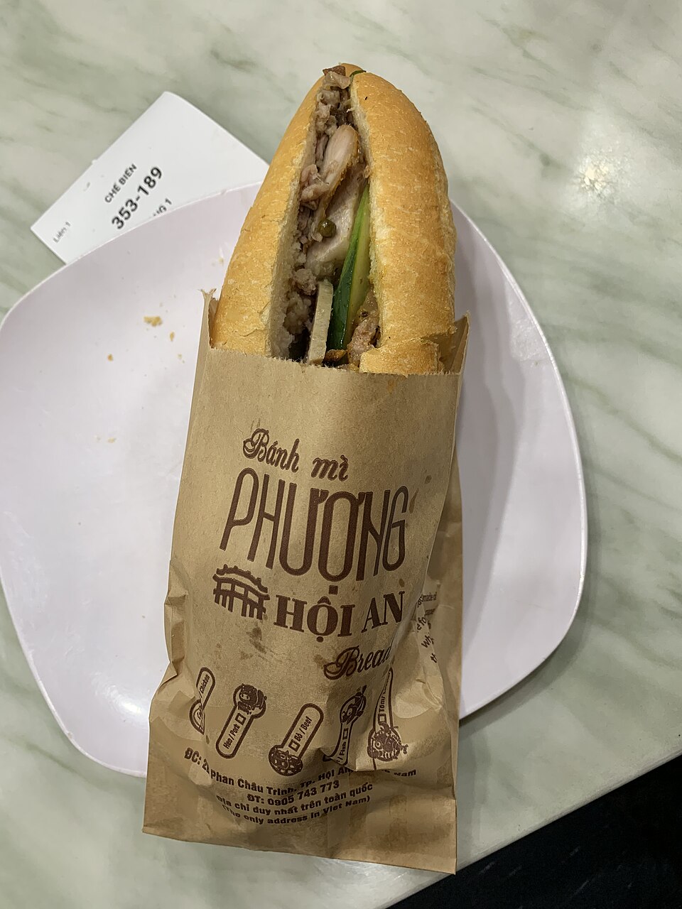 A loaf split lengthwise sits on a plate inside a brown paper bag printed 'Bánh mì Phượng Hội An'. The loaf is filled with meat, cucumber and pork roll, and the loaf tapers to a point at the top.