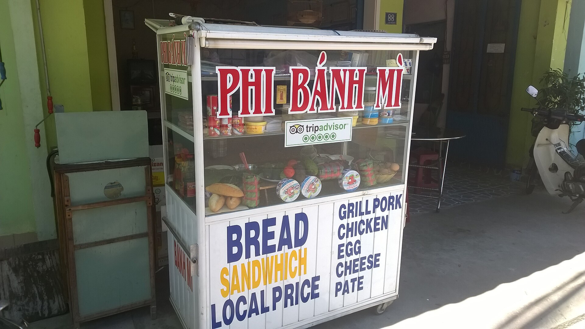A glass-fronted bread stall by the roadside with a white sign in red letters reading 'PHI BÁNH MÌ'. Its front carries English lettering about bread, sandwiches and local price, and a list of fillings, and inside are a few loaves, cheese boxes, tins and vegetables.