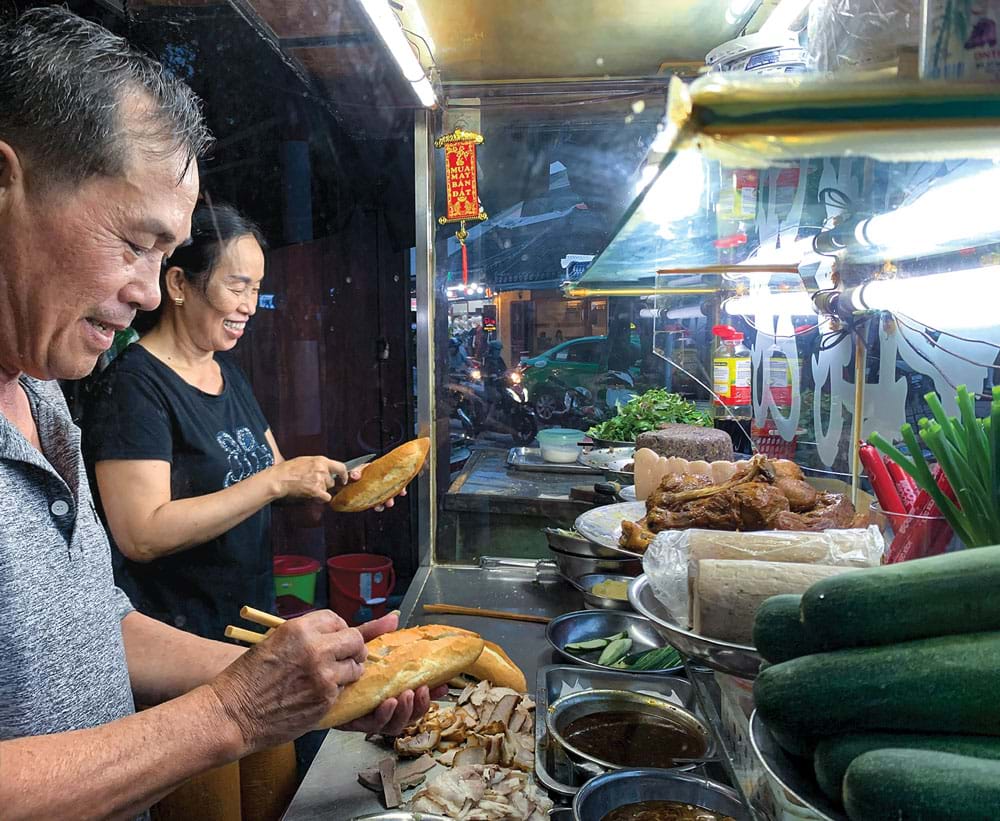 Two people stand behind a glass-fronted bread stall at a street corner at night: a man in a grey shirt slits a loaf, a woman in black behind him holds another, and the counter holds sliced xíu pork, roast poultry, pork roll, cucumbers, spring onions and chillies.