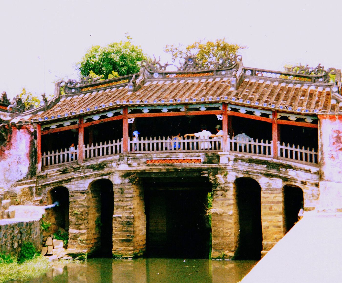 The bridge photographed in 2003 from the creek: weathered stone piers, faded red columns, moss on the tiles.