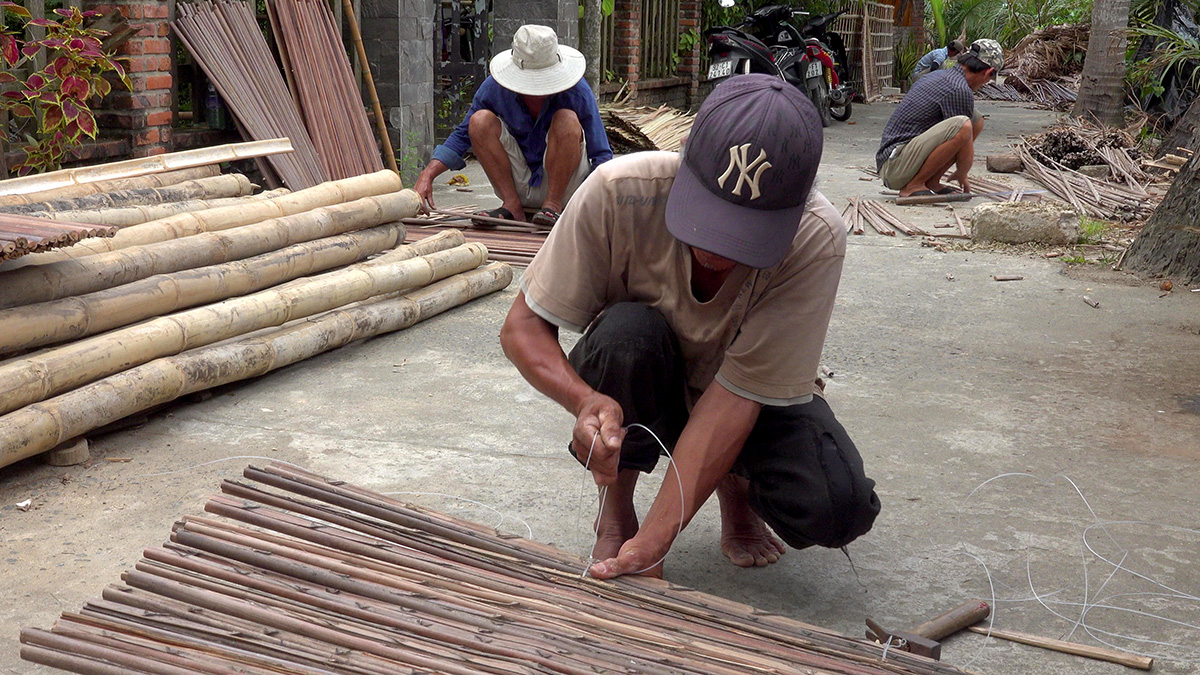 A worker crouches tying cord onto a woven bamboo-strip panel laid on a concrete yard, split-bamboo bundles piled around him, two more workers bent over their own panels further back in the same yard.