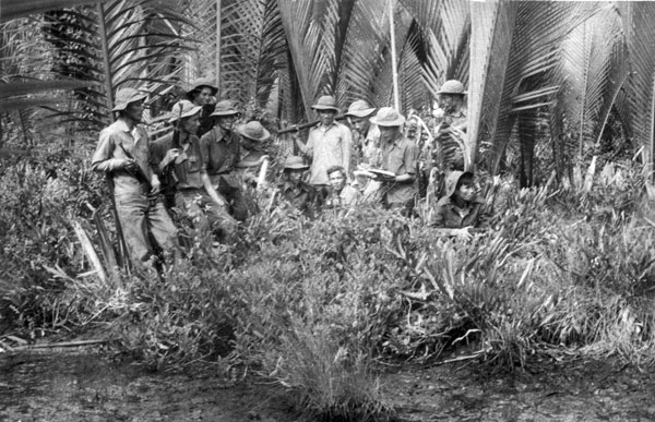 Black-and-white photograph of around ten men in soft field caps, one with a rifle slung over his shoulder, standing and crouching among nipa-palm fronds taller than head height, one man at the centre holding a sheet of paper the others are looking at.