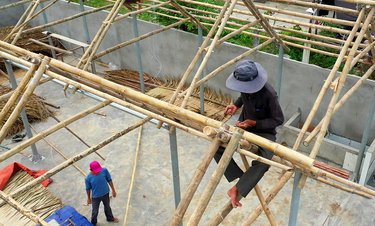 Overhead view of a bamboo frame under construction on a concrete slab, bamboo poles crossing into triangular trusses, a hatted worker seated on the frame tying a joint, dried coconut leaves piled below.