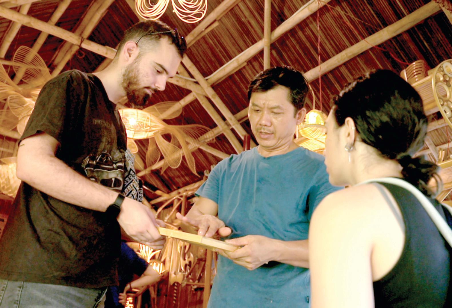 Inside a coconut-leaf-roofed space, woven bamboo lamps and fish shapes hanging from the ceiling, a craftsman holding out a small bamboo item for a foreign visitor to look at, another visitor watching beside them.