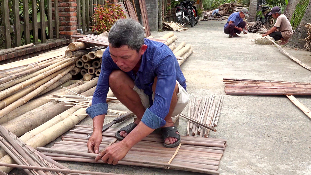 A yard in Cẩm Thanh with several workers weaving bamboo panels at once on the concrete floor, whole bamboo poles and split strips piled around the yard, a motorbike parked behind them.