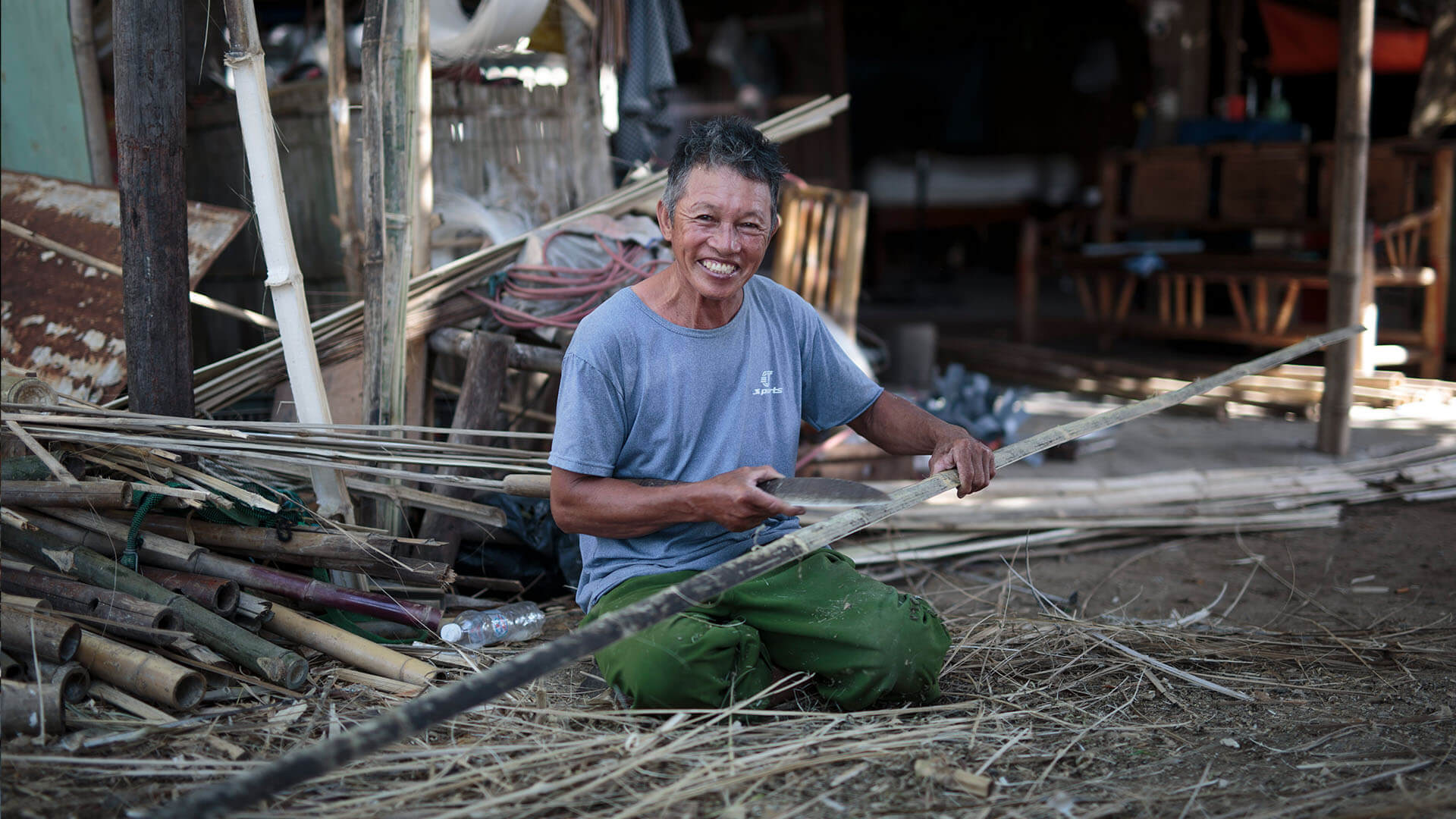 Portrait of an older worker, greying hair, smiling broadly, both hands holding a small knife as he splits a long bamboo pole, whole bamboo culms piled in the yard behind him.