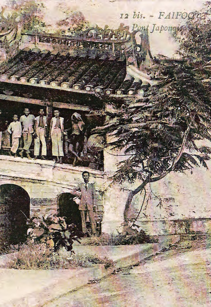 A tinted postcard captioned at the top “12 bis. – FAIFOO / Pont Japonais”. The western end of the bridge seen at an angle: tiled roof and decorated ridge, five people standing on the deck under the roof, a man in a European suit at the foot of the stone abutment with its arched opening, and a large tree hanging into half the frame.