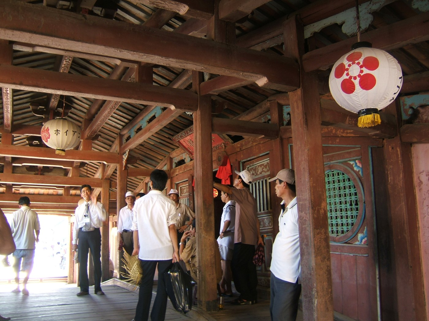 Inside the covered bridge looking along the passage: dark timber posts and the exposed roof frame overhead, raised plank walkways down both sides of the central lane, a round window with green lattice in the right-hand wall, two paper lanterns hanging above, and several people walking through or standing to look.