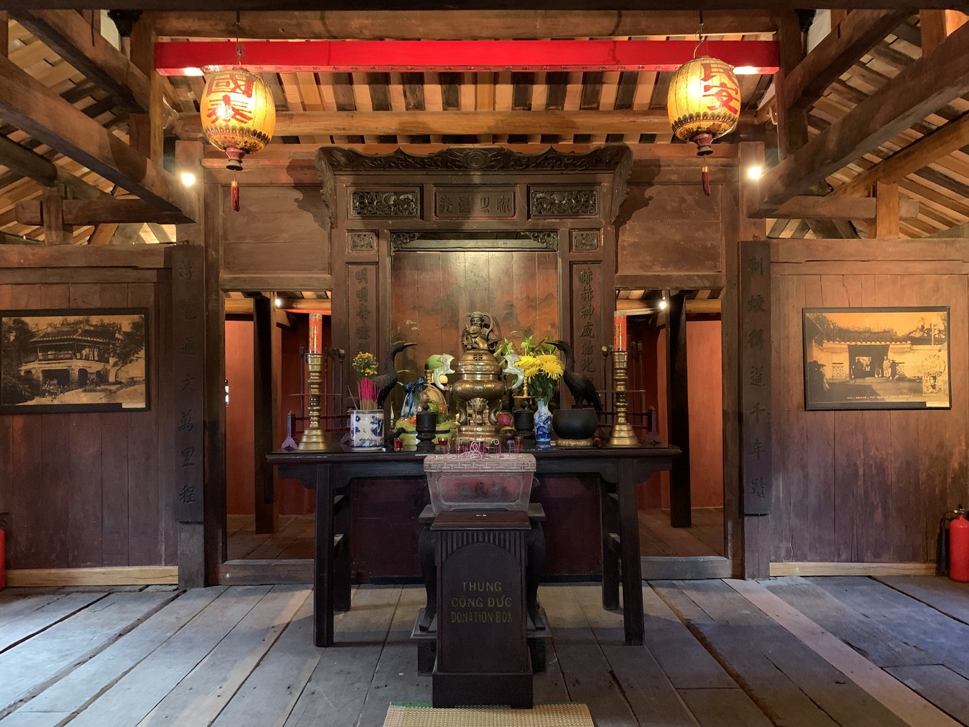 The shrine seen head on: a carved wooden shrine frame with a small dark figure inside it, and in front a wooden table set with a bronze incense burner, bronze candlesticks, blue and white porcelain vases and two vases of fresh flowers. A red-painted beam runs above, a paper lantern lettered in Chinese characters hangs on either side, two framed old photographs of the bridge hang on the timber walls, and a donation box stands on the plank floor.