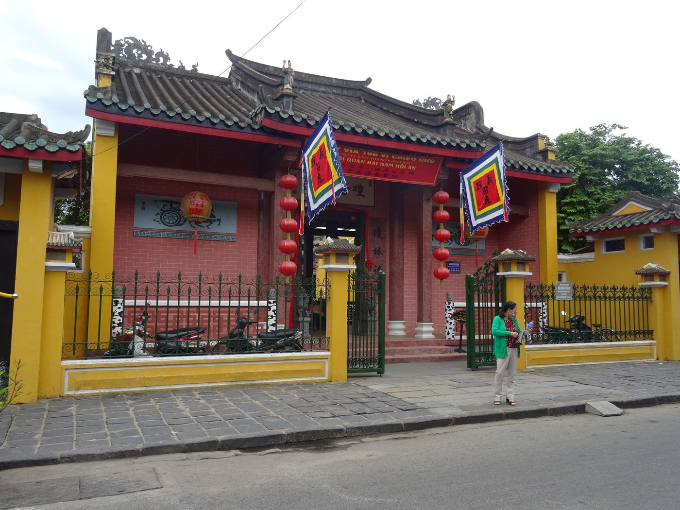 The front of the Hainan hall in Hội An on a feast day, yellow boundary wall, red-pink façade, a red banner strung across the front, two festival flags in Chinese characters standing either side of the main door, strings of red lanterns.