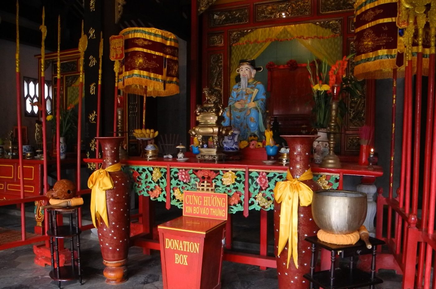 A side altar inside the Quan Công temple: a red lacquered table carved with coloured flowers, a seated figure in blue robes and a winged cap in its shrine, a bronze censer, two embroidered canopies above, ceremonial halberds ranked either side, and a small sign asking that incense money go in the box.