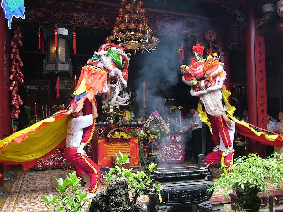 Two lion-dance teams lift their lion heads before an altar wreathed in incense smoke, an old chandelier and red couplets behind, potted plants set around the courtyard.