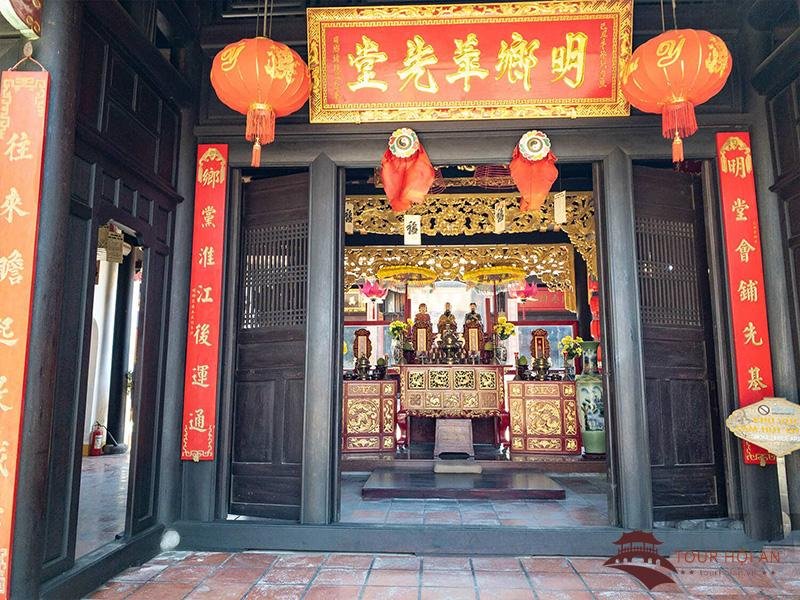A view through the central doorway into the main hall: a red-and-gilt board with four Chinese characters hanging above, a pair of red couplets running down the door frame, red lanterns on both sides, and a gilt-carved shrine deep inside holding three small seated figures on a dais.