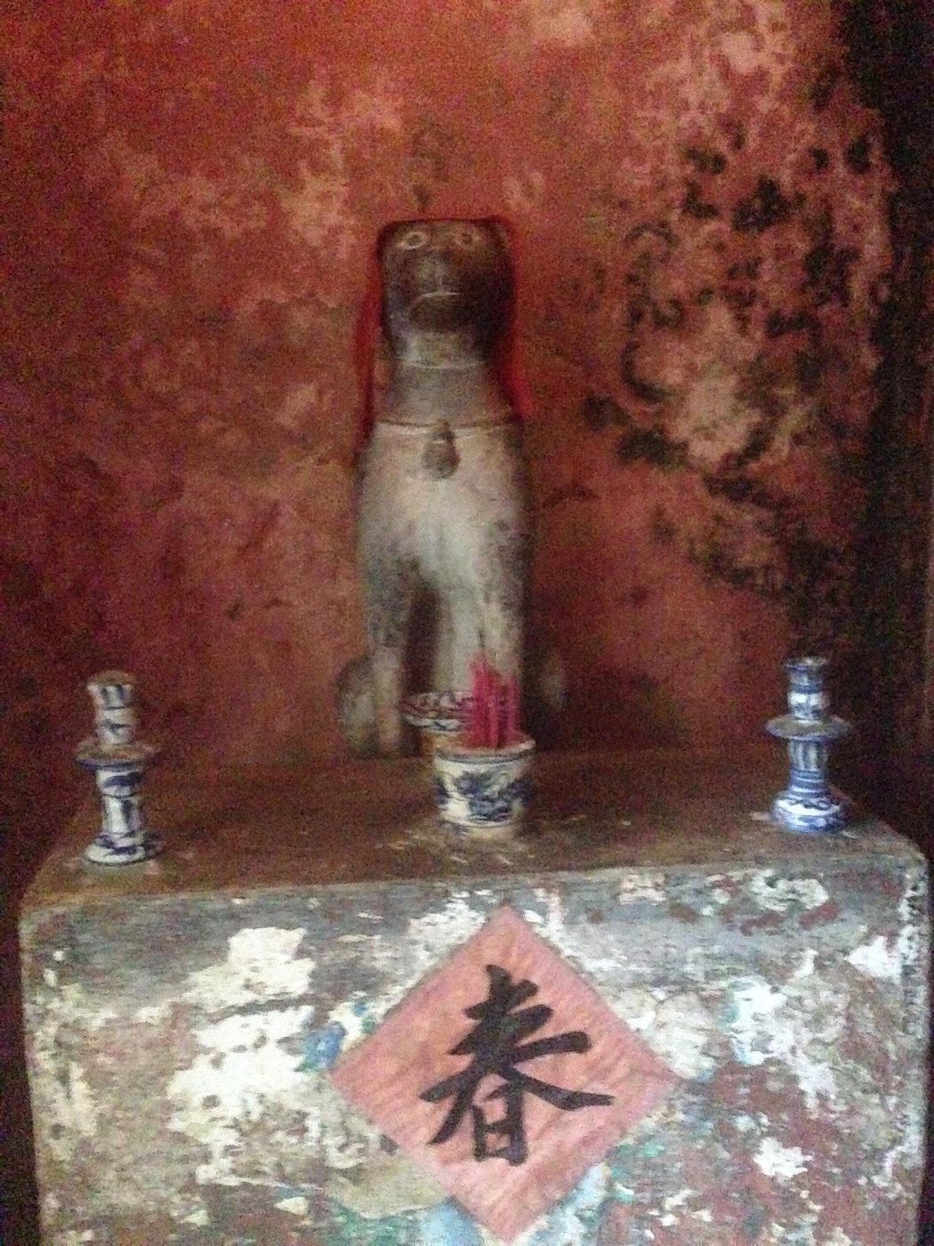 The wooden dog guardian at one end of the bridge, on a worn altar with incense and a red paper charm.