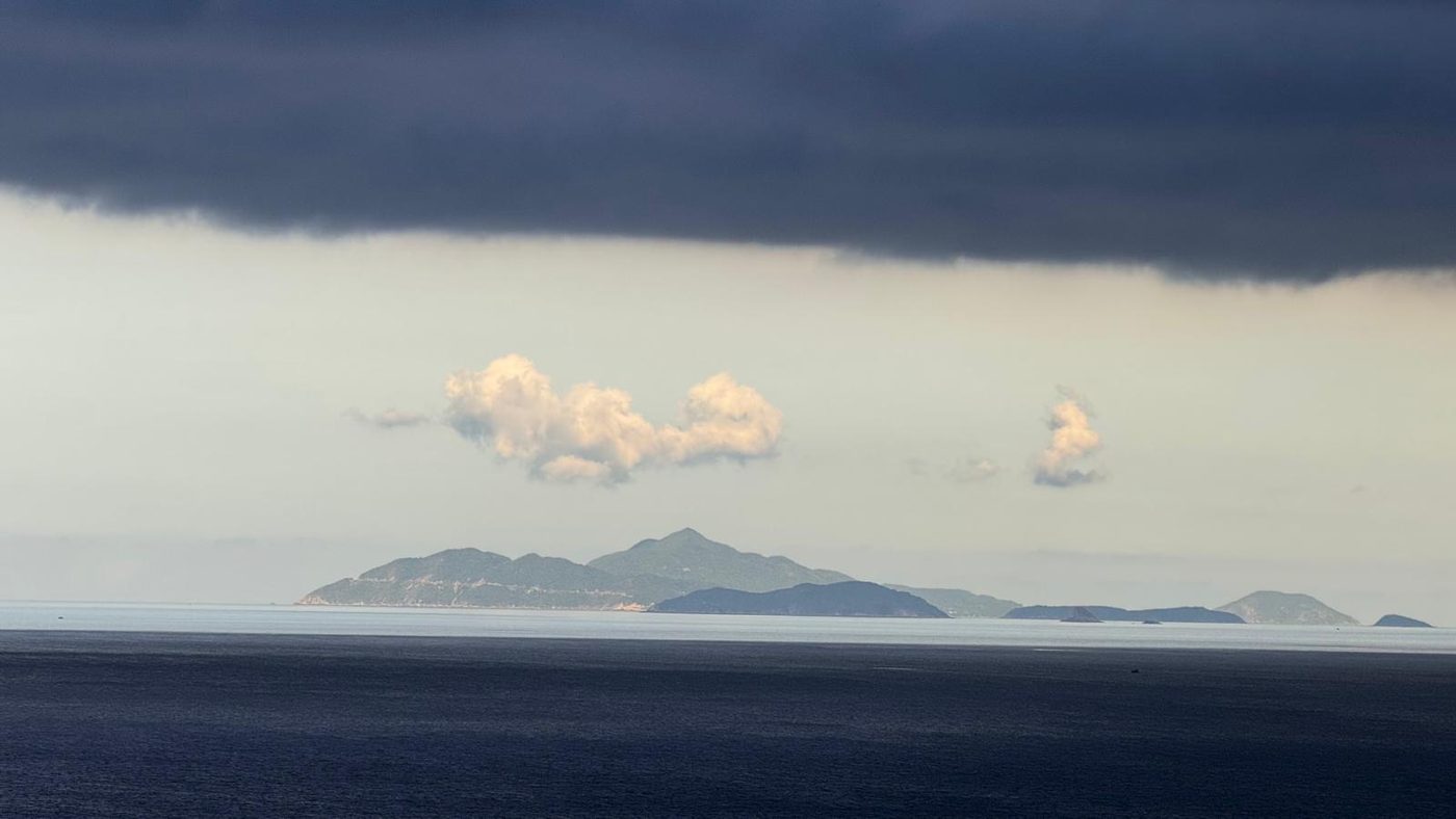 Cù Lao Chàm seen from the mainland across dark water, the green ridge of the island lying along the horizon beneath a low bank of storm cloud.