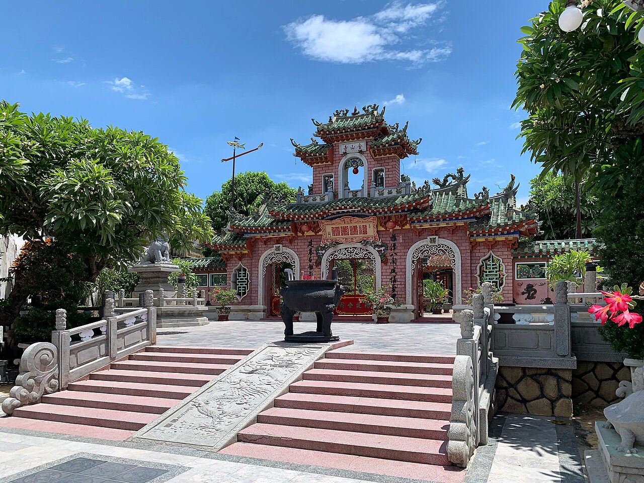 The pink triple gate of the Fujian Assembly Hall in Hội An under a blue sky, with green glazed roof tiles, a bronze censer in the forecourt and a carved dragon ramp on the steps.