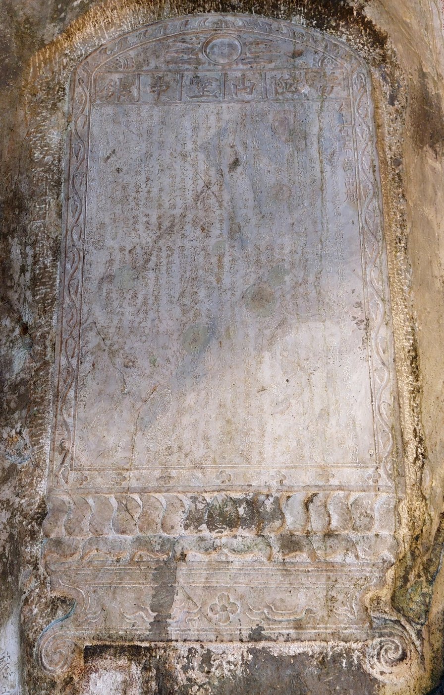 A close view of the stone cut into the cave wall: an arched frame with a sun-and-moon motif at the top, a horizontal line of four characters as the title, many columns of vertical text below, and a wave motif at the foot.