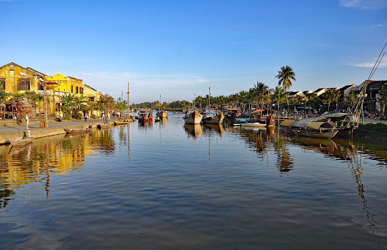 The Thu Bồn river at Hội An in late afternoon: yellow-walled shophouses along the left bank, wooden boats moored in rows on the right, still water reflecting both.