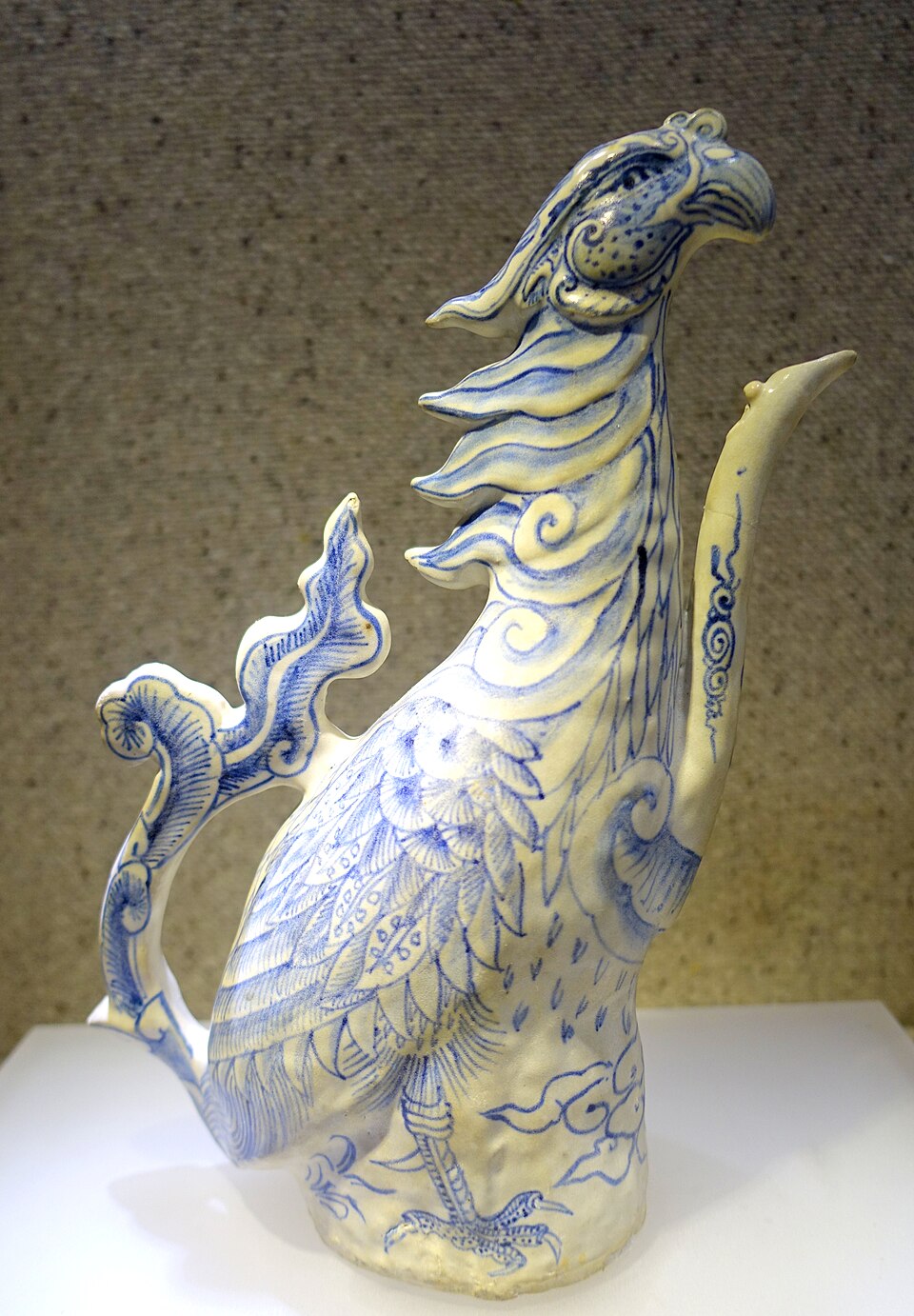A blue-and-white ceramic ewer shaped as a standing phoenix, painted with feathers, scrolling clouds and a flowering branch, displayed in a museum case.
