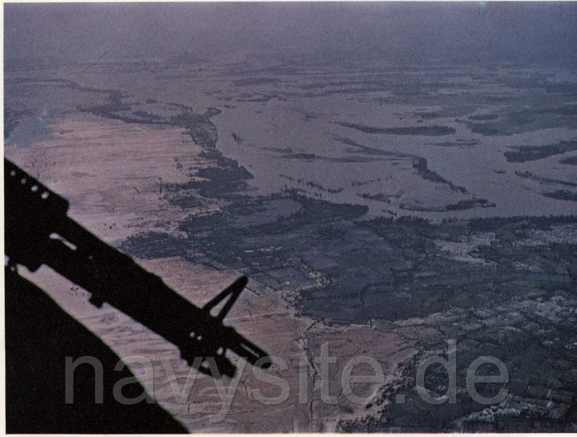 A colour photograph taken past the door gun of a helicopter: the coastal plain under water, river channels and paddy bunds showing only as faint lines.