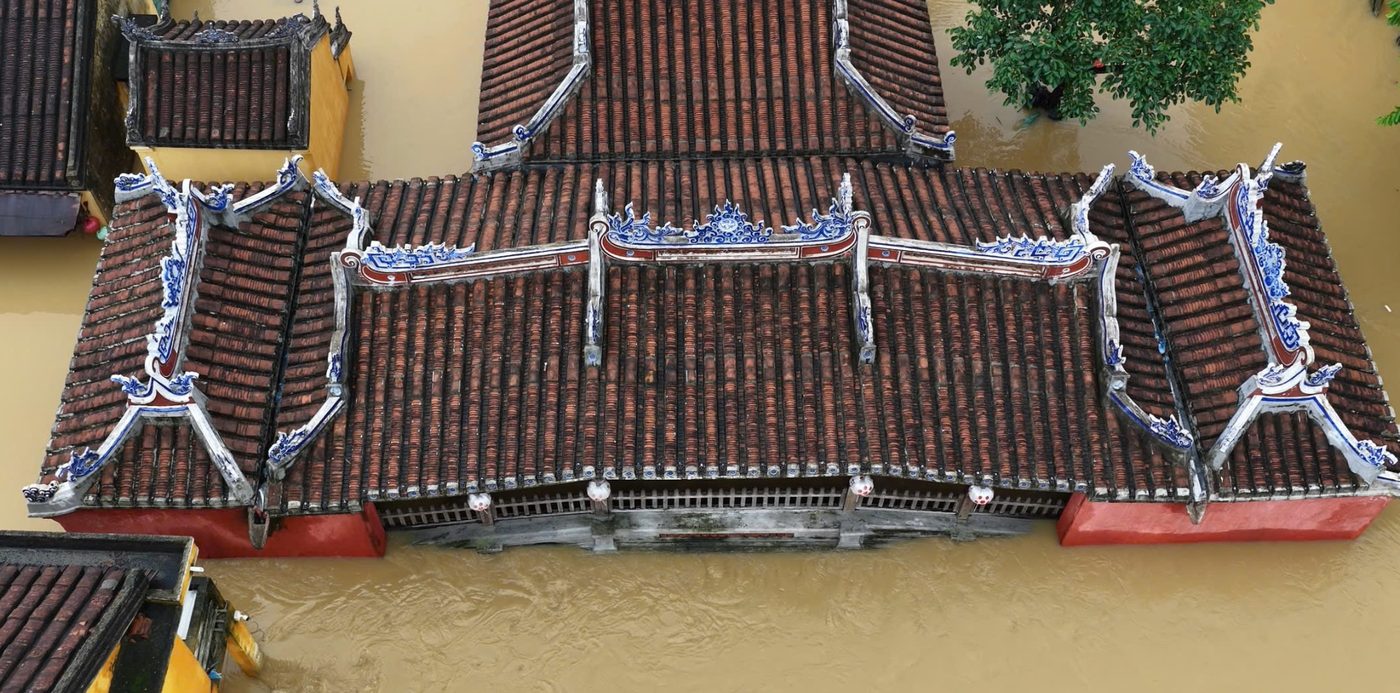 Straight down onto the covered bridge from the air: the tiled roof and its blue-and-white ridge ornaments stand clear, and the brown water is up against the eaves on both sides.