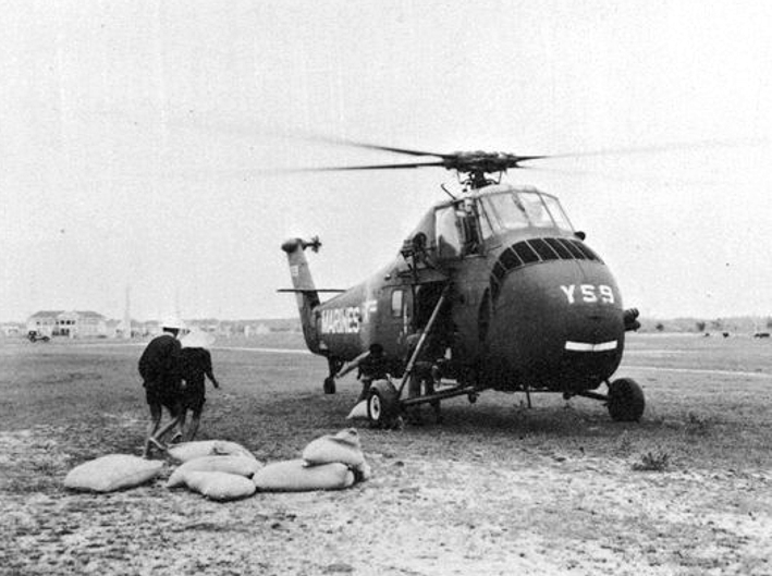 A Sikorsky UH-34D marked MARINES, side number YS9, stands on a grass airstrip; two men carry the last sacks of relief supplies clear of the aircraft.