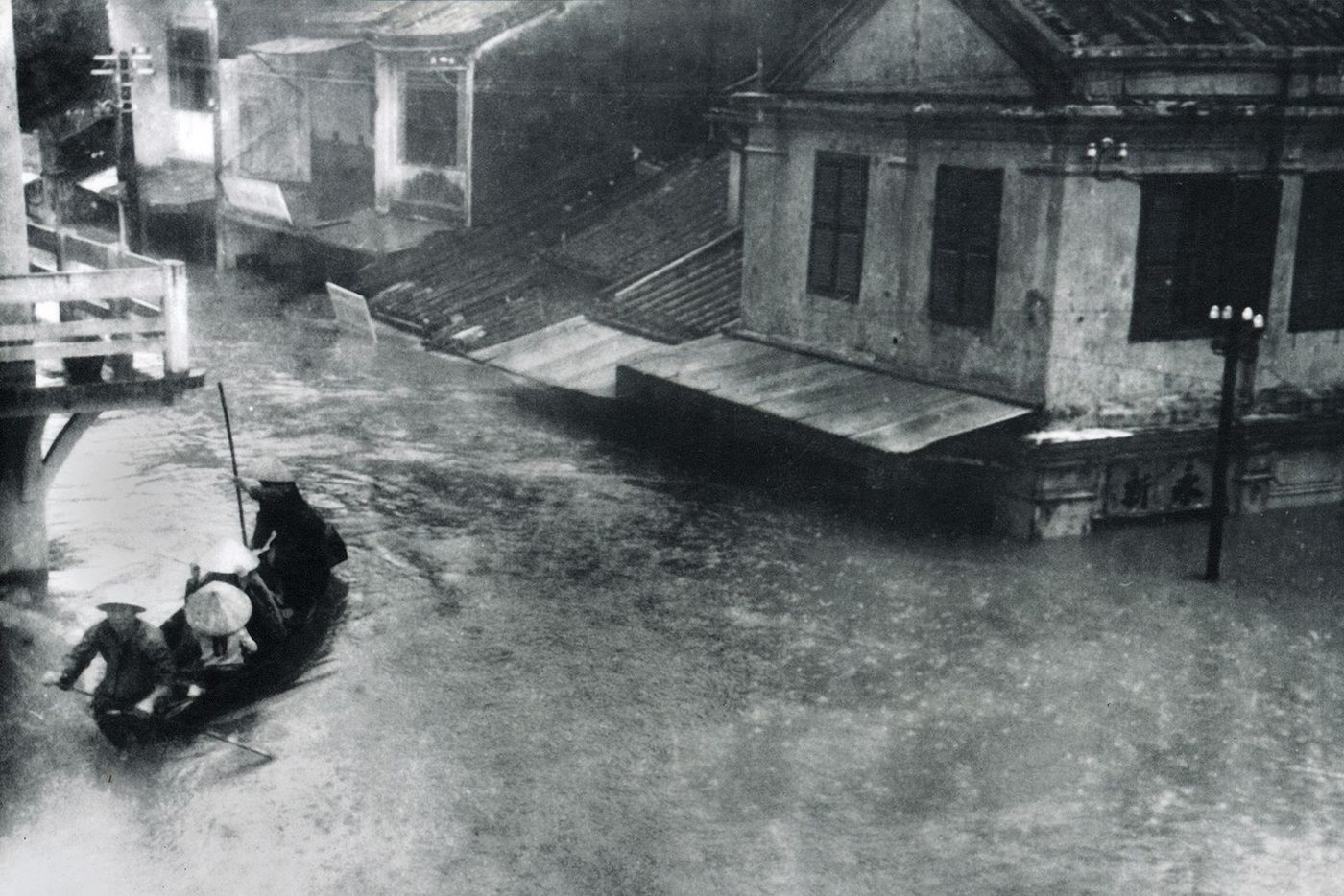 Seen from an upper floor: the ground floor of a corner house is entirely under water, only the upstairs shutters still dry, while three people in conical hats pole a small boat past the foot of a bridge.