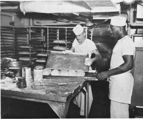 Two of the ship's bakers at a steel table below deck, sliding a tray of dough into a rack of ovens.