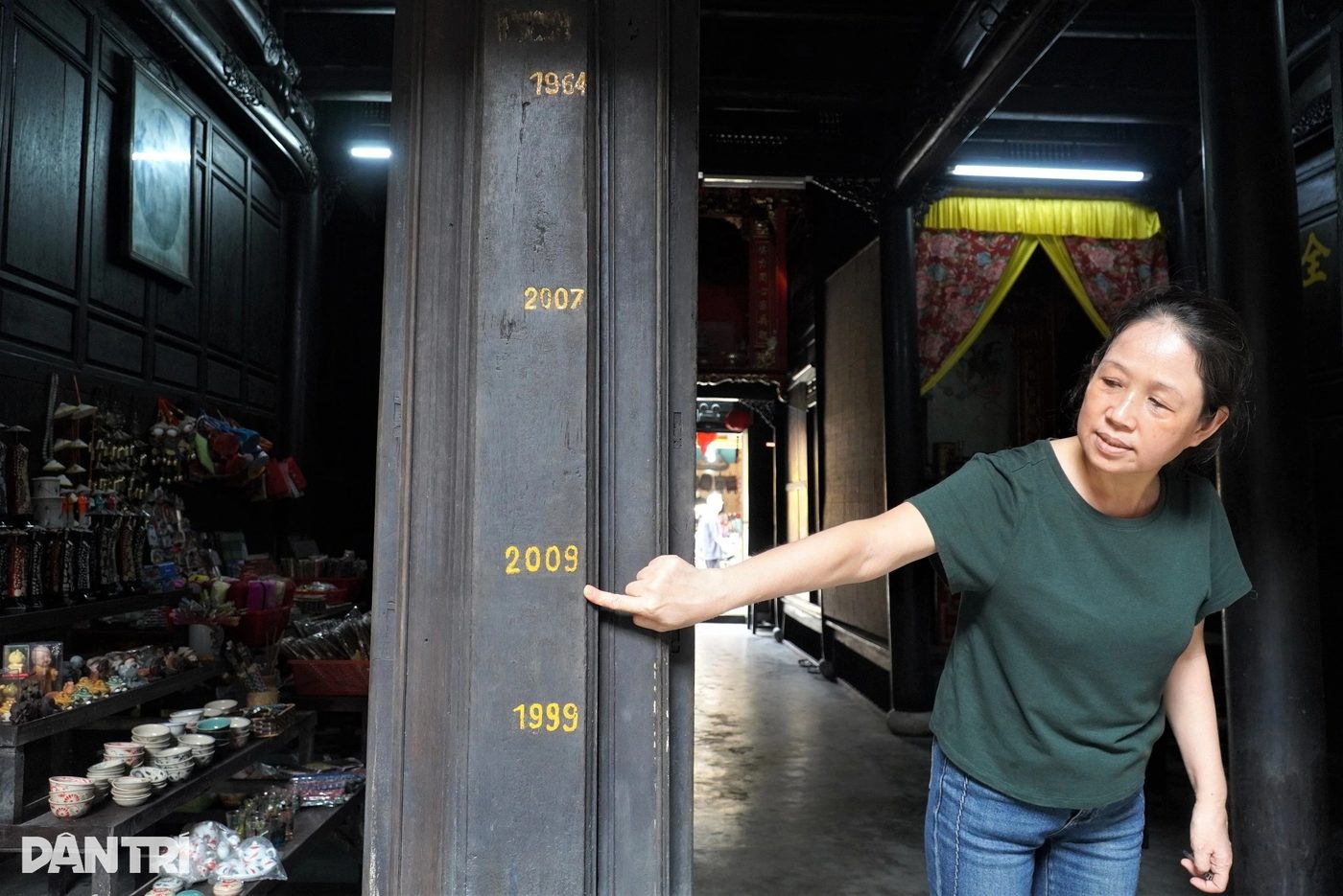 A wooden column inside the Quân Thắng house painted with gold flood-year marks — 1964 at the top, then 2007, 2009 and 1999 — with Diệp Ái Phương reaching up to touch the 2009 line.