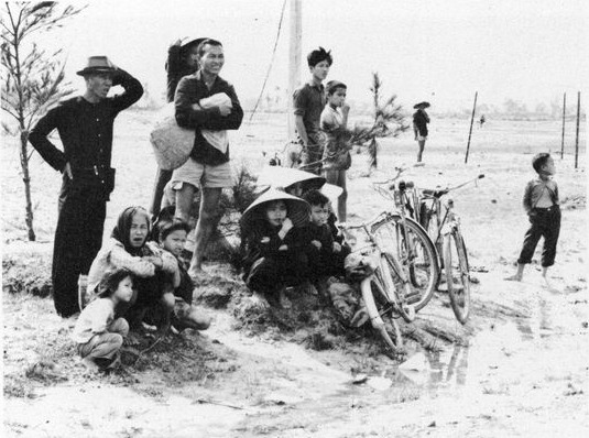 Villagers waiting at the edge of the Tam Kỳ landing field: adults standing, children sitting on the sand, bicycles laid over in the grass.
