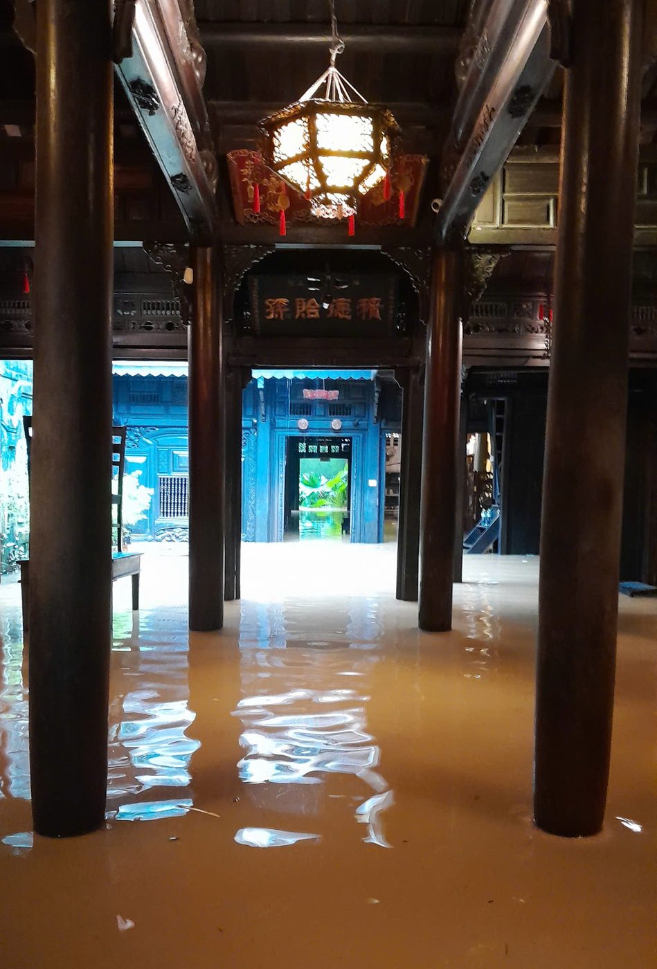 Inside the Tấn Ký house with floodwater standing across the tiled floor between the ironwood columns, a lantern lit above, the blue-painted rooms beyond half under water.