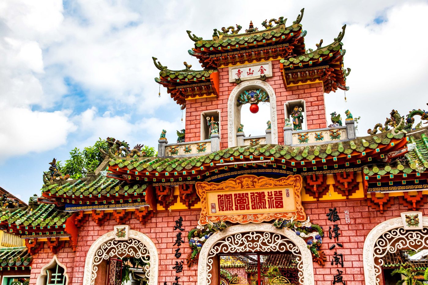 The upper tier of the rebuilt gate, a vertical plaque reading Kim Sơn Tự set into red brickwork below layered green-tiled eaves, a red ball ornament hanging in the archway beneath it.
