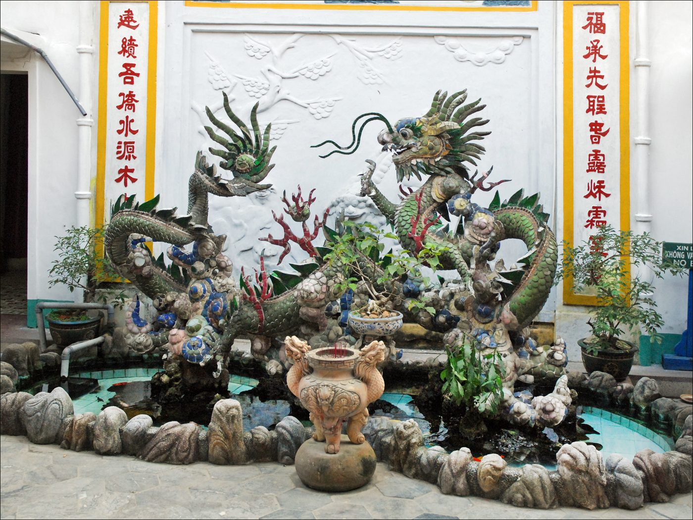 A ceramic-and-glass mosaic dragon coiling over a small courtyard pond between the main and rear sanctuaries, a stone incense burner in front, red-lettered couplets on the wall behind.