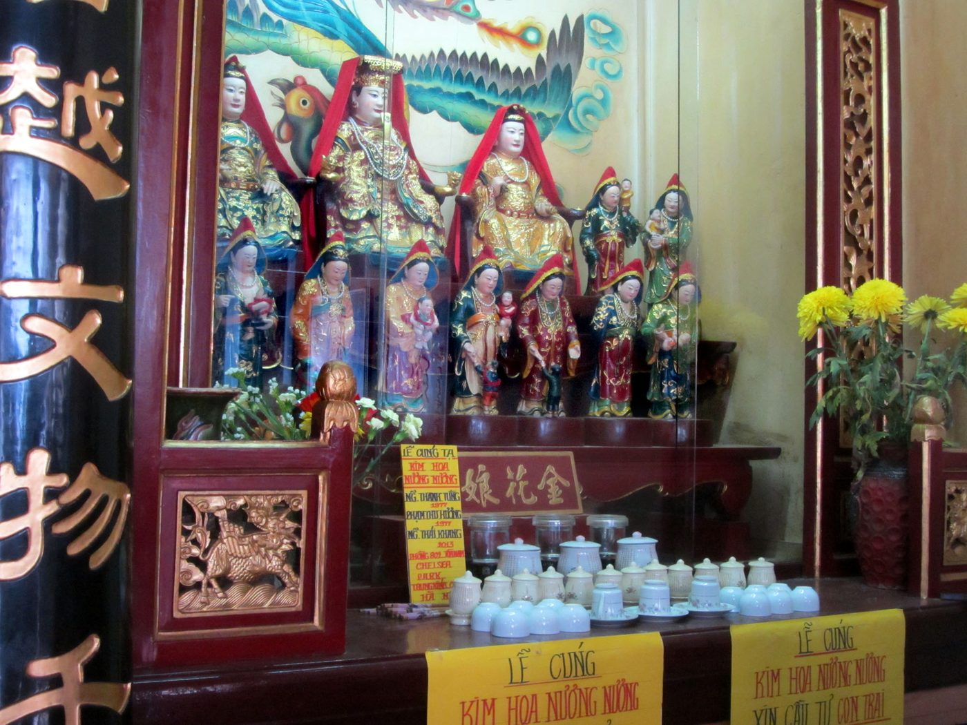 An altar case holding gilded seated goddess figures on an upper tier and a row of standing attendant figures below, several cradling infants, marigolds and offering cups on the table in front, a sign at the base reading in Vietnamese and Chinese characters for the goddess Kim Hoa Nương Nương.