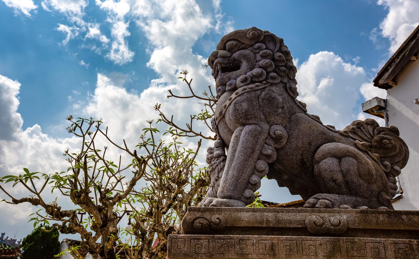 A weathered stone guardian lion on a carved pedestal at the hall's entrance, seen from below against a bright sky, a frangipani branch beside it.