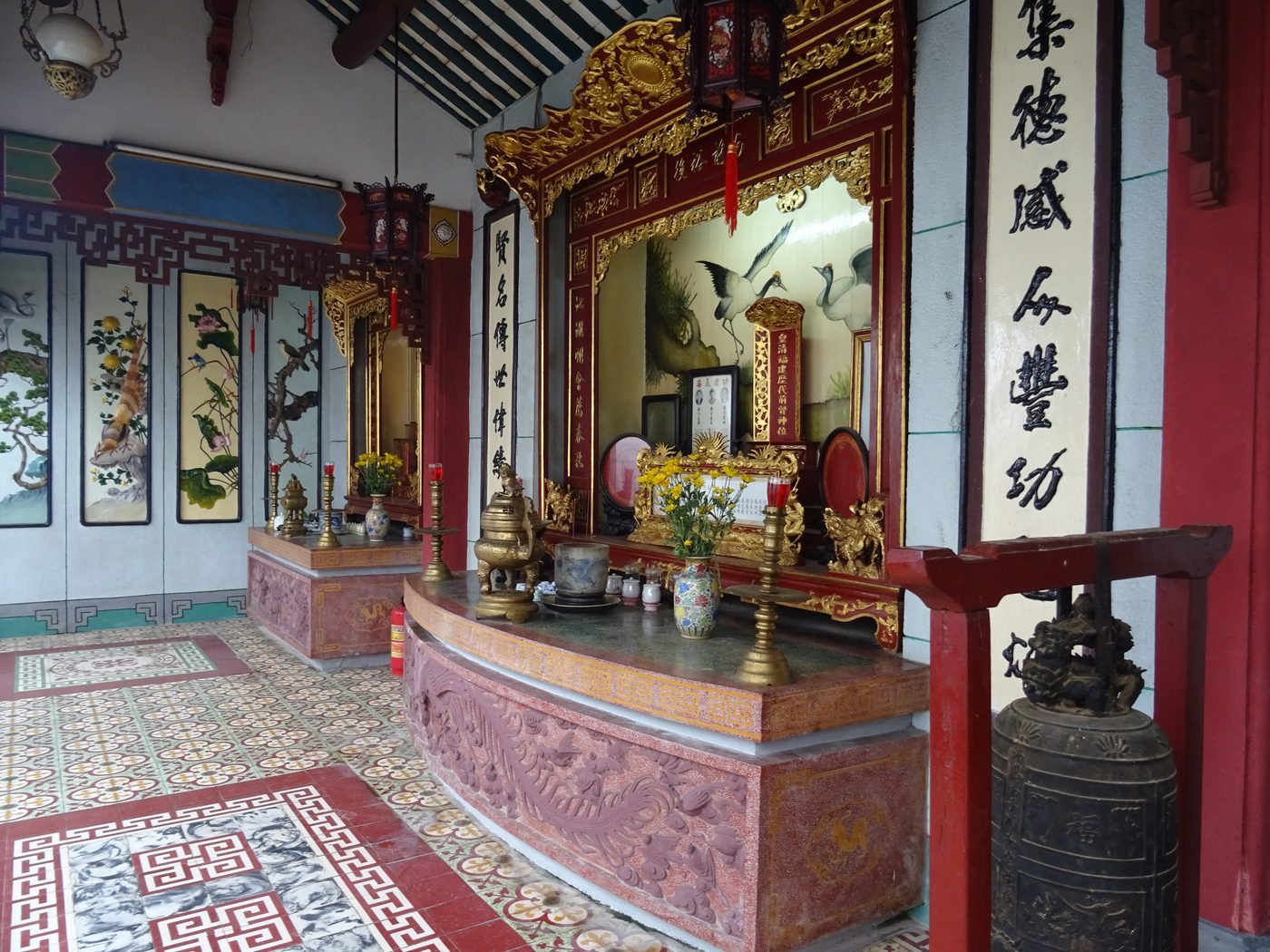 A side hall inside the assembly hall with tiled floor patterns, an ancestral altar bearing gilded couplets, an old bronze bell resting on a red stand in the foreground.