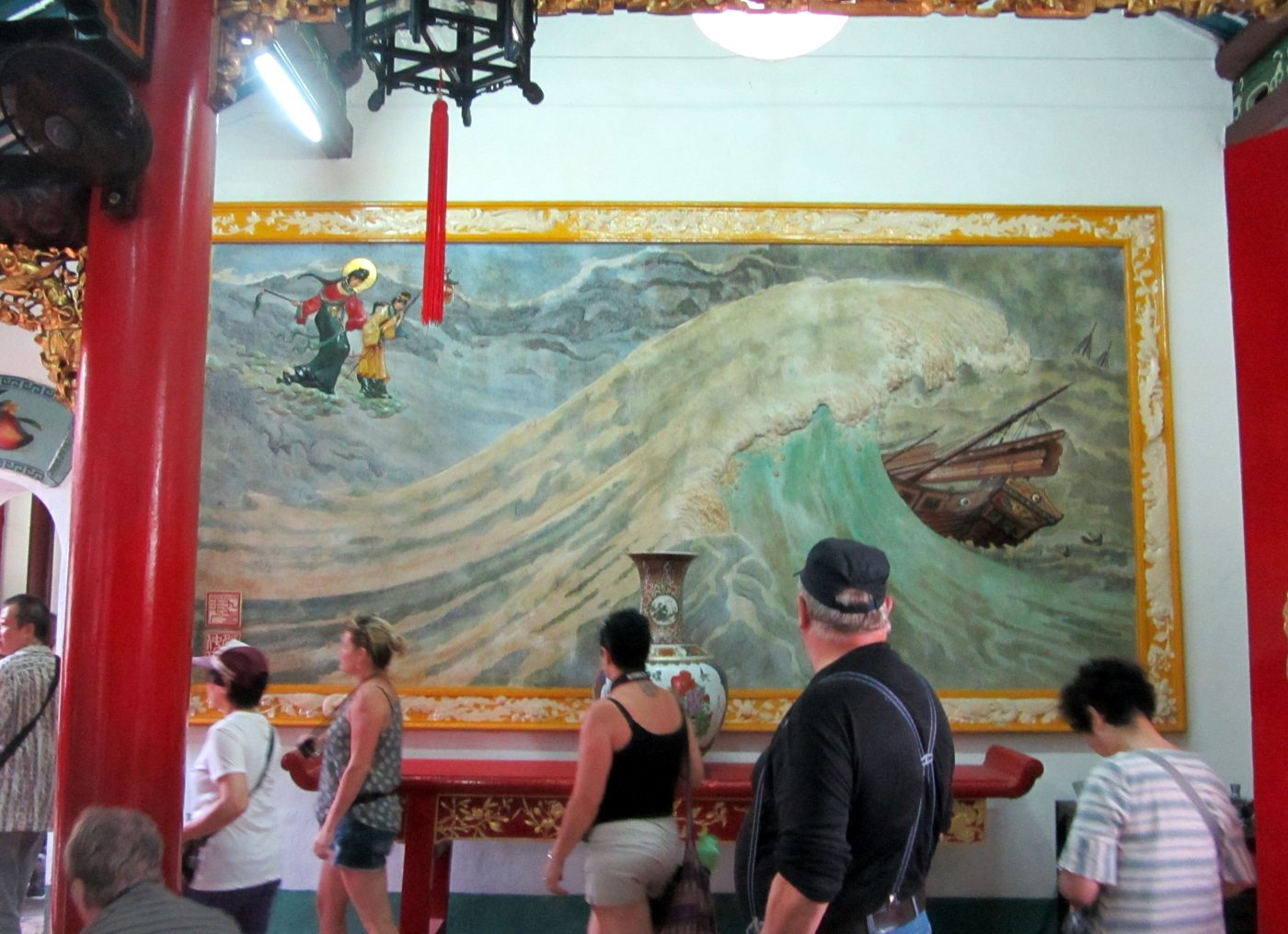 A wall painting inside the hall showing Thiên Hậu and an attendant walking on stormy waves toward a foundering wooden junk, viewed by a group of visitors.