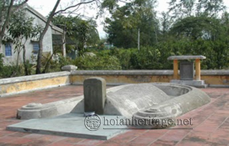 The restored grave enclosure: red brick paving, a yellow-washed wall around it, a stone stele standing before the saddle-shaped mound, and a small tiled shelter behind to the right.