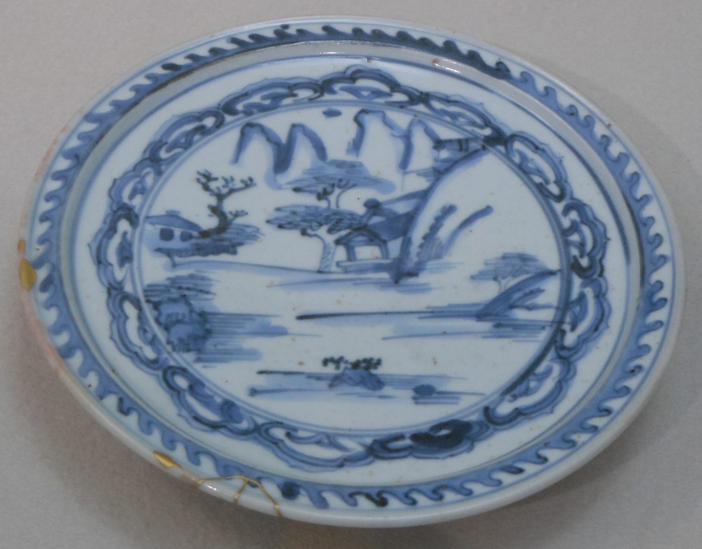 A round porcelain dish, white glaze painted in cobalt blue: hills, trees and a roof by water in the well, a band of cloud scrolls around the rim and a saw-tooth border outside it. A repair in gold runs across the edge.