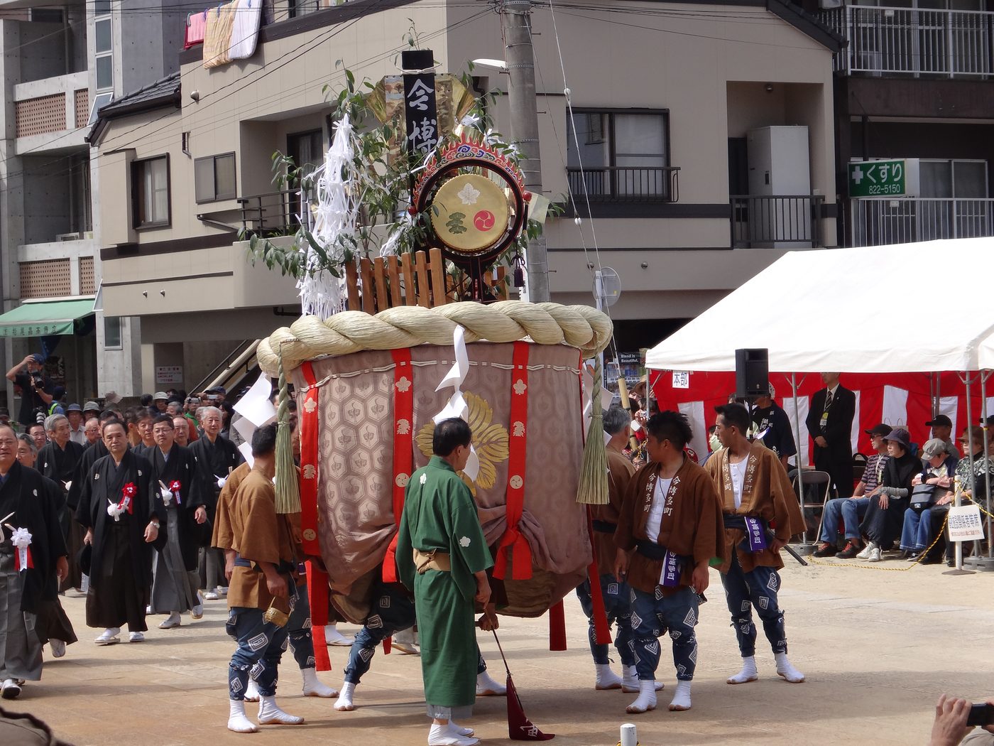 A large canopied float bound with a plaited rope, a drum and green branches set on top, carried through the street by men in brown and green coats with a crowd watching on both sides.