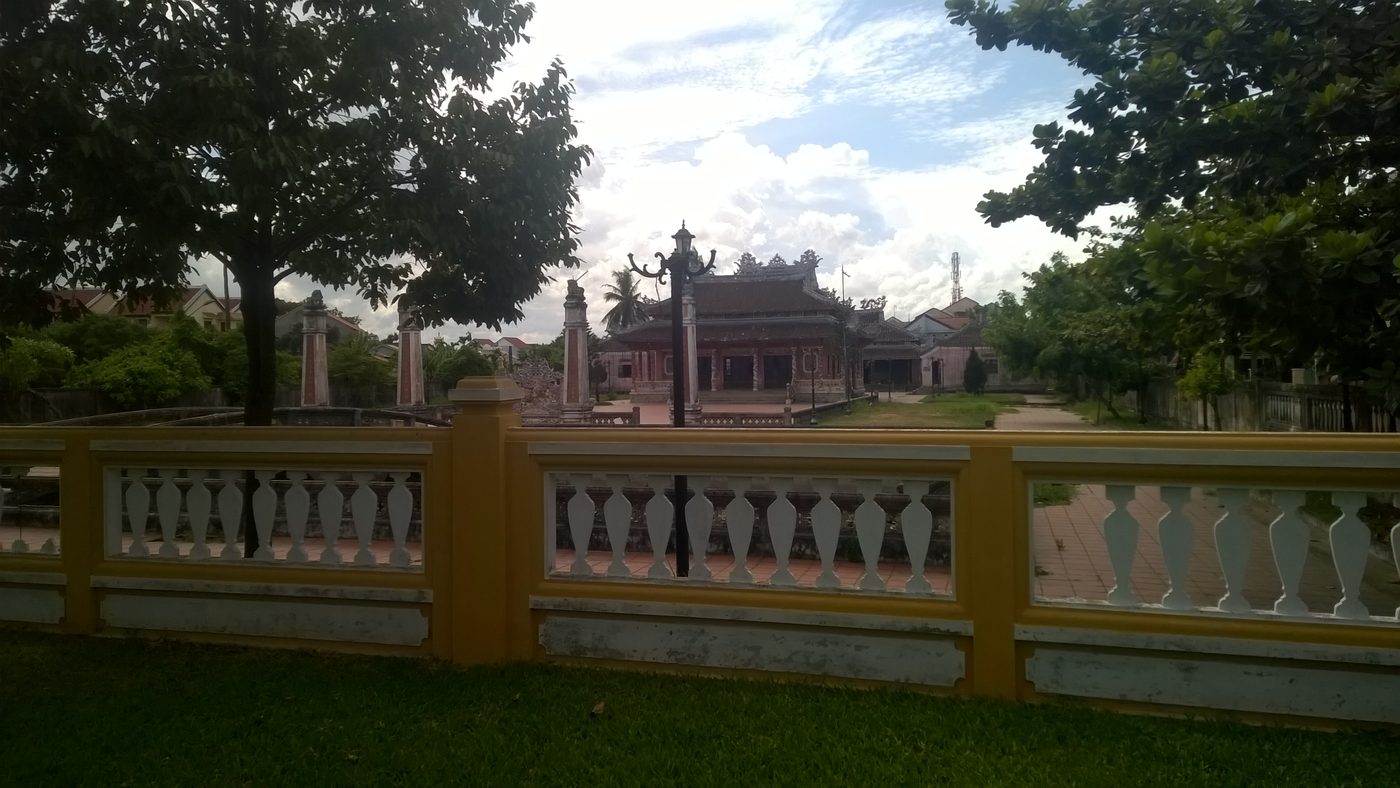 A tiled shrine with curved ridges and pillars flanking its yard, seen across a yellow and white balustrade, trees on both sides and a clouded sky.