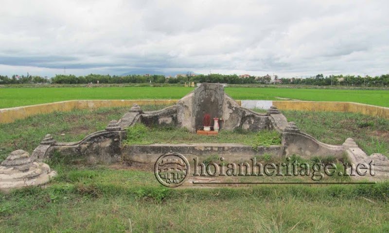 A small grave enclosure amid green paddy fields, a low curved wall on each side, and a single stone stele at the centre with an incense bowl and a water offering.