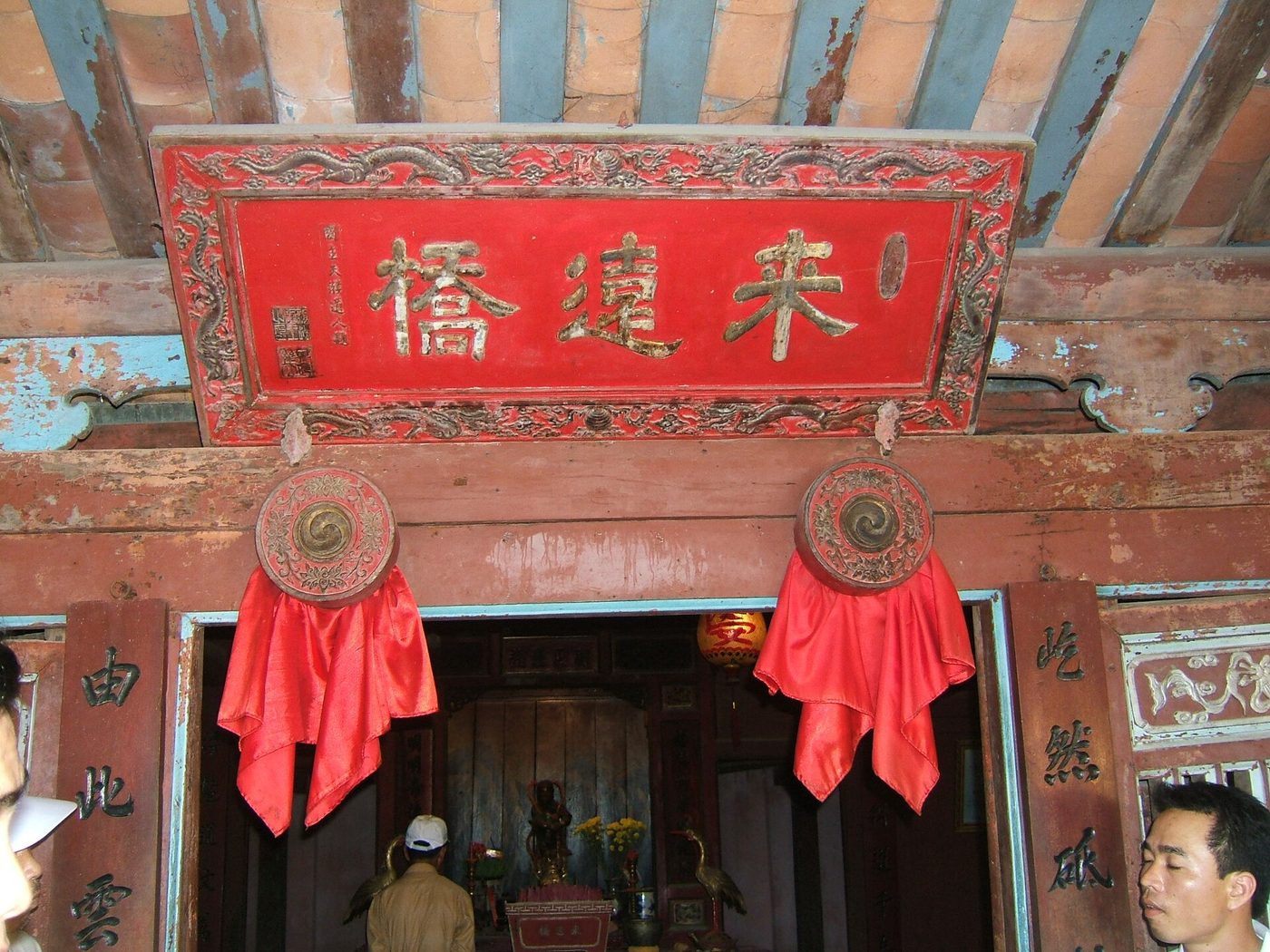 The red-lacquered board carved 來遠橋 in gilt characters, hanging above the shrine doorway between two red cloth rosettes.