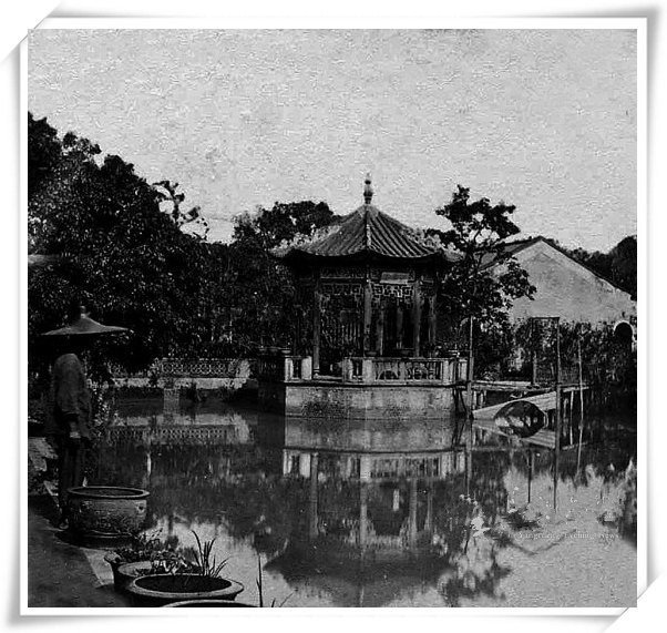 An old black-and-white photograph: a tiled pavilion standing over a still pond, its reflection in the water, dense trees around it, a figure in a conical hat standing at the bank in the foreground.