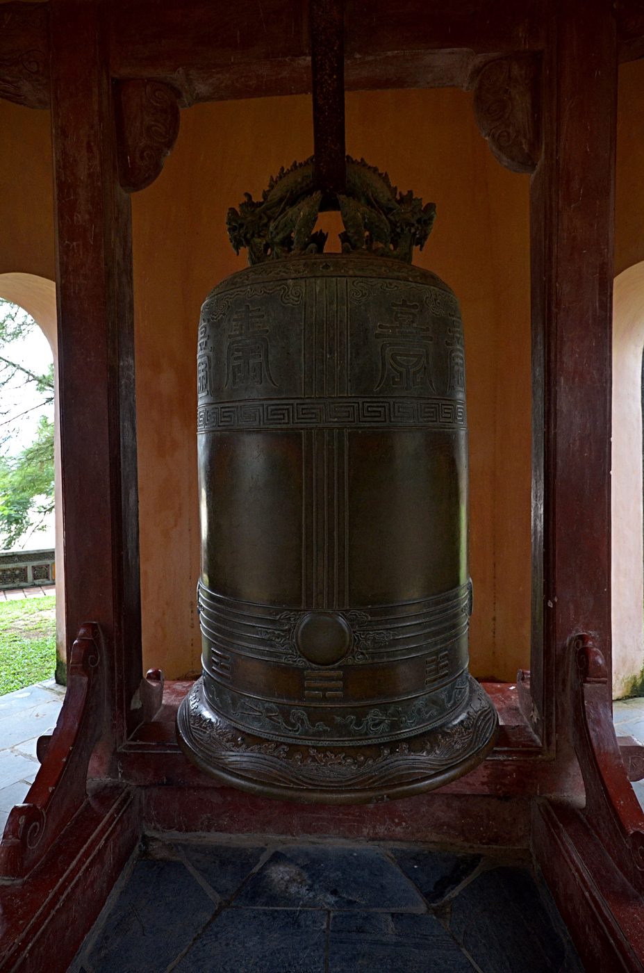 A large bronze bell hung in a yellow-walled pavilion: dragons cast on the crown, Chinese characters in roundels on the body, a band of key fret around the shoulder, trigrams lower down and a band of waves at the foot.