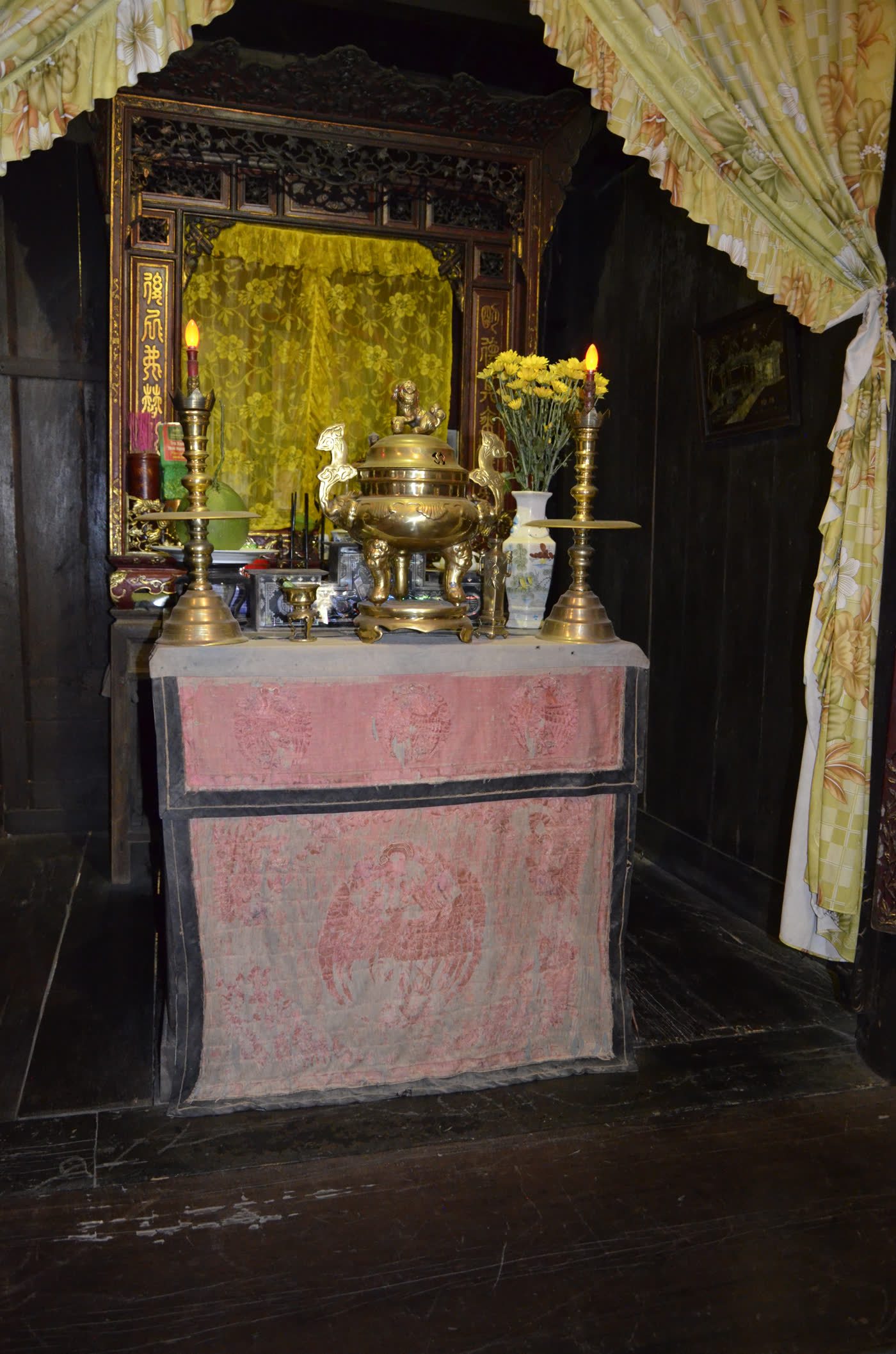 A family ancestral altar inside Tấn Ký house, a bronze incense burner and a pair of tall candlesticks in front of a gilt shrine board, marigolds in a vase beside it, a pink embroidered altar cloth draped over the front.